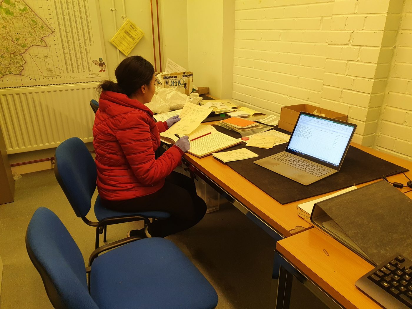 A woman sitting down wearing a red jacket sitting on a blue chair. The woman is holding an archival document and recording the data in a book. The woman is a room with other archival documents and a laptop.
