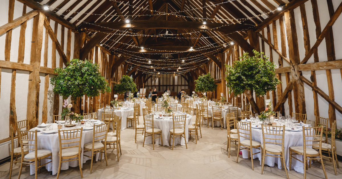 Wedding set up in a old barn - round tables with white tables cloths, wooden gold chairs, fairy light hung up between the wooden beams of the barn