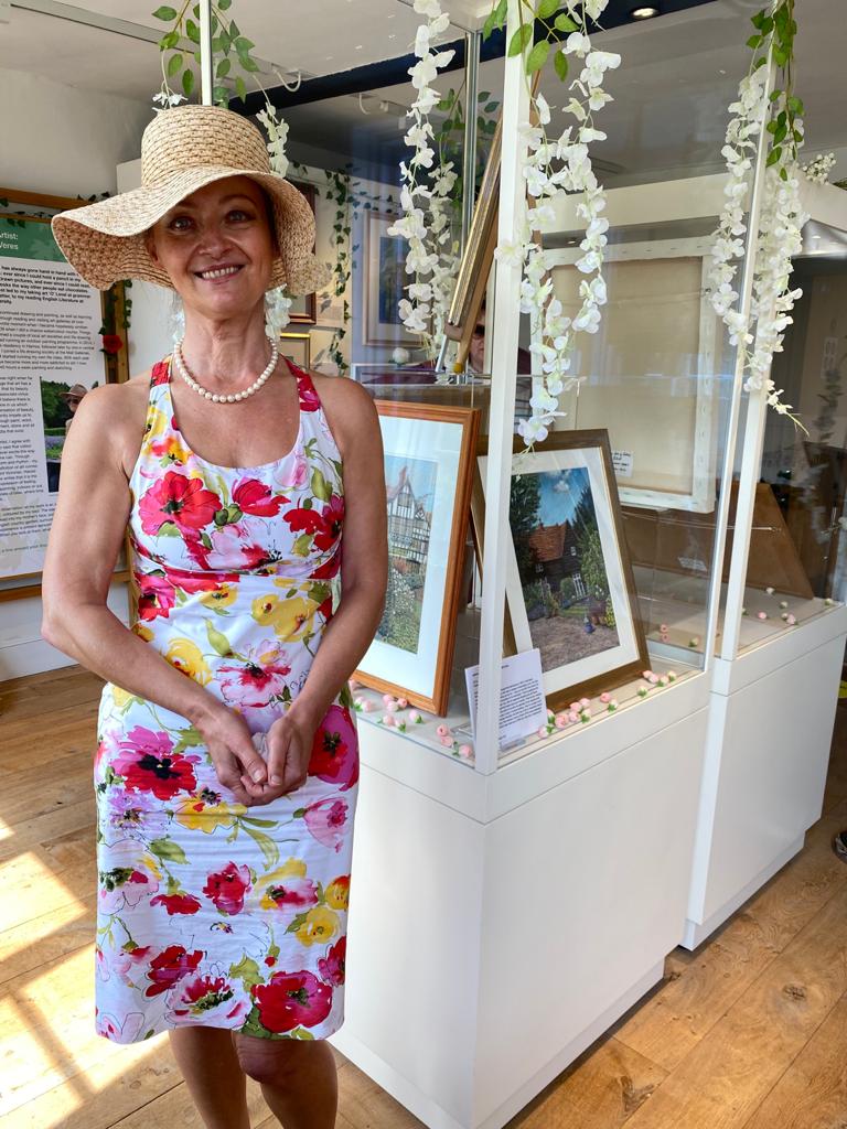 Alexandra Veres inside the exhibition room standing next to the exhibition she curated. Alexandra is wearing a large straw summer hat and wearing a summer dress with pink, yellow and red flowers on it.